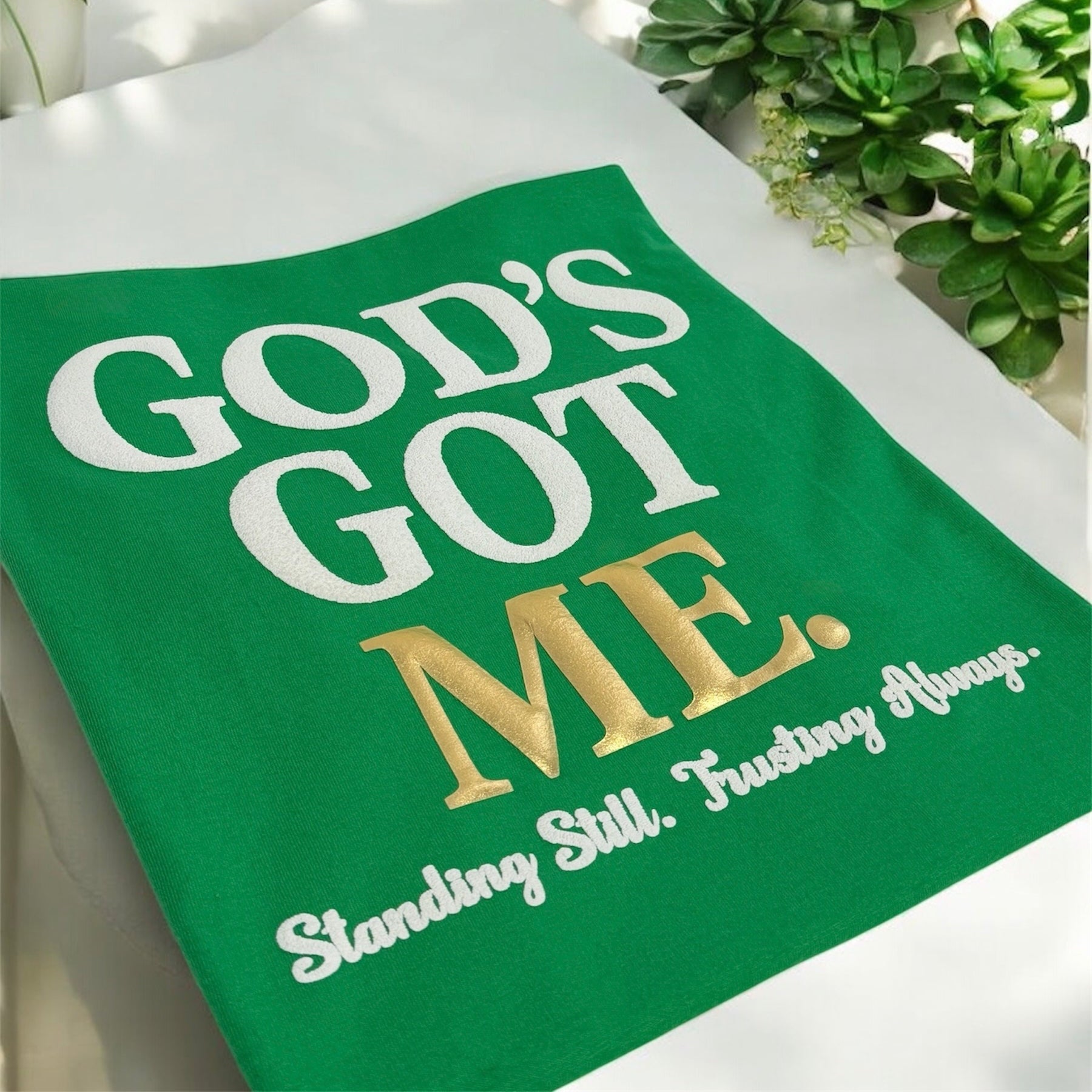 Green towel with 'God's Got Me' text on a white surface with plants in the background