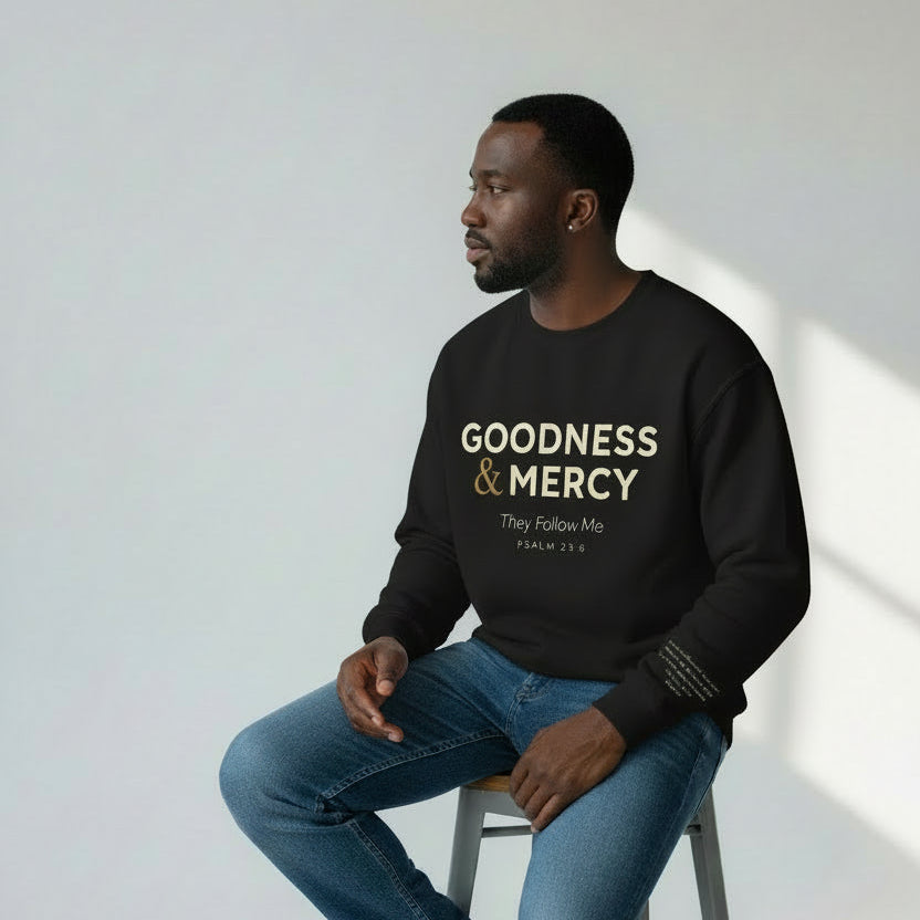 Man wearing a black sweatshirt with 'Goodness & Mercy' text, sitting on a stool against a white wall.