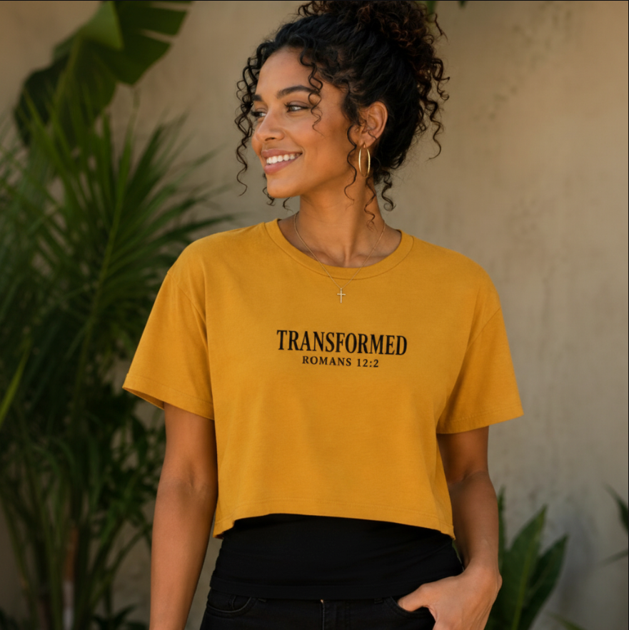 A woman wearing a mustard yellow cropped t-shirt with 'TRANSFORMED ROMANS 12:2' printed in black on the chest, paired with black pants, set against a warm neutral wall with green plants.