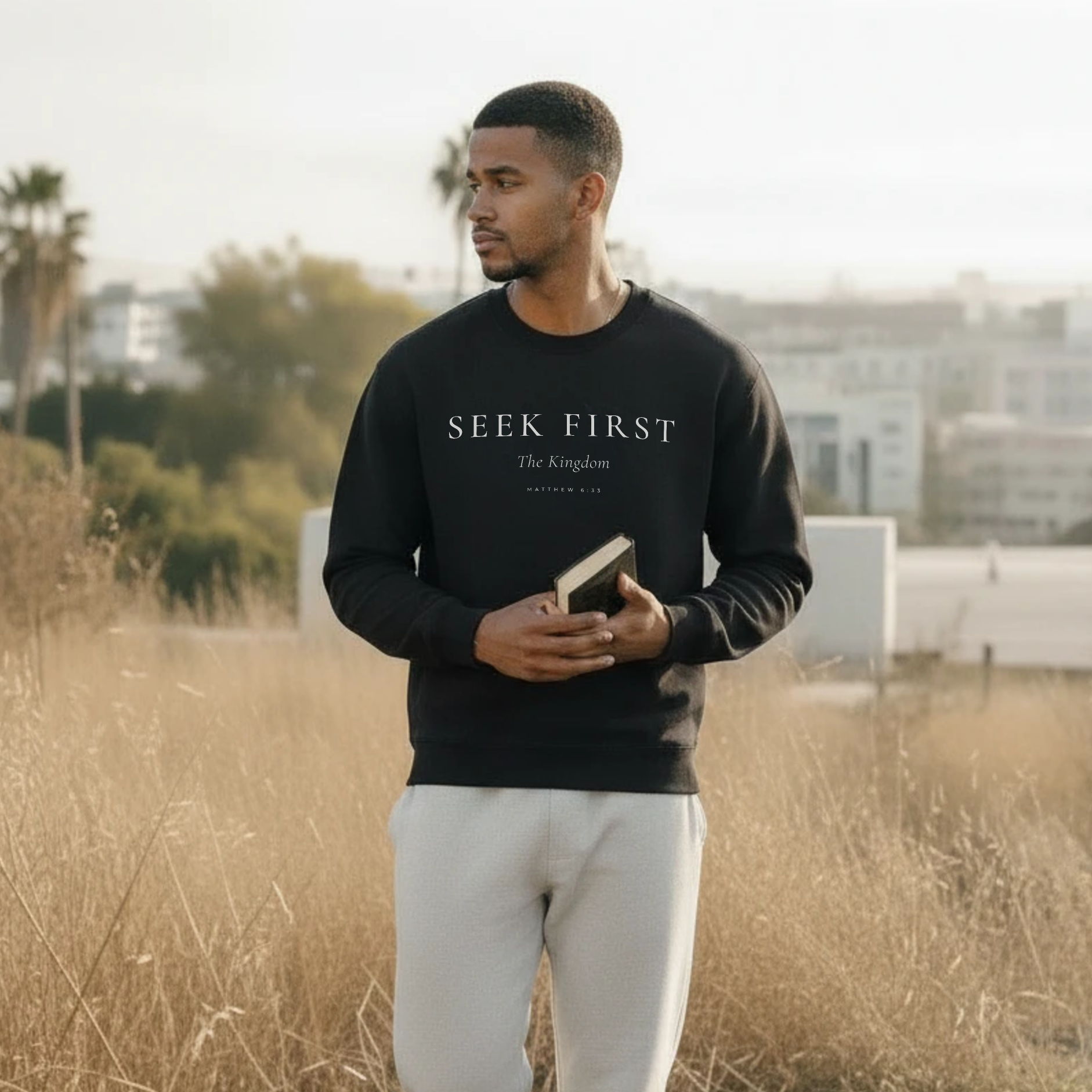 Man wearing a black sweatshirt with 'Seek First' text, holding a book, standing in a field.