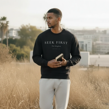 Man wearing a black sweatshirt with 'Seek First' text, holding a book, standing in a field.