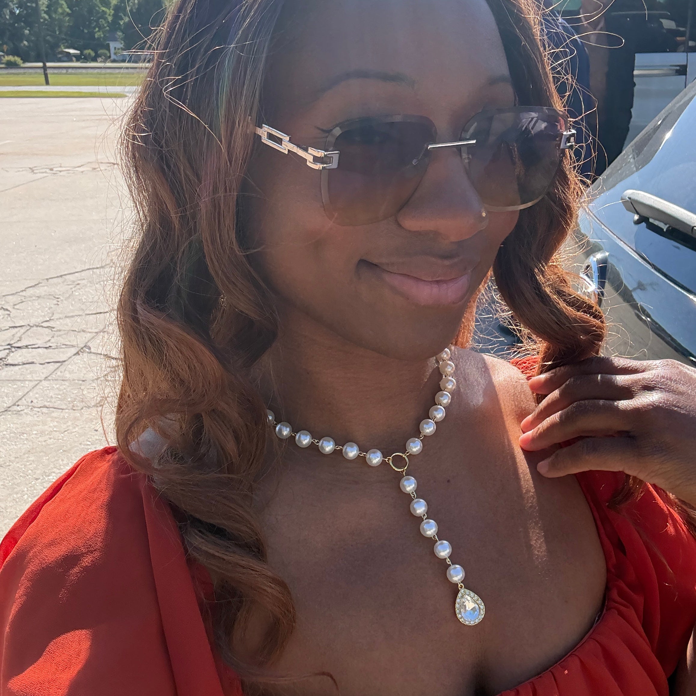 Woman wearing sunglasses and a red dress with a pearl necklace outdoors.