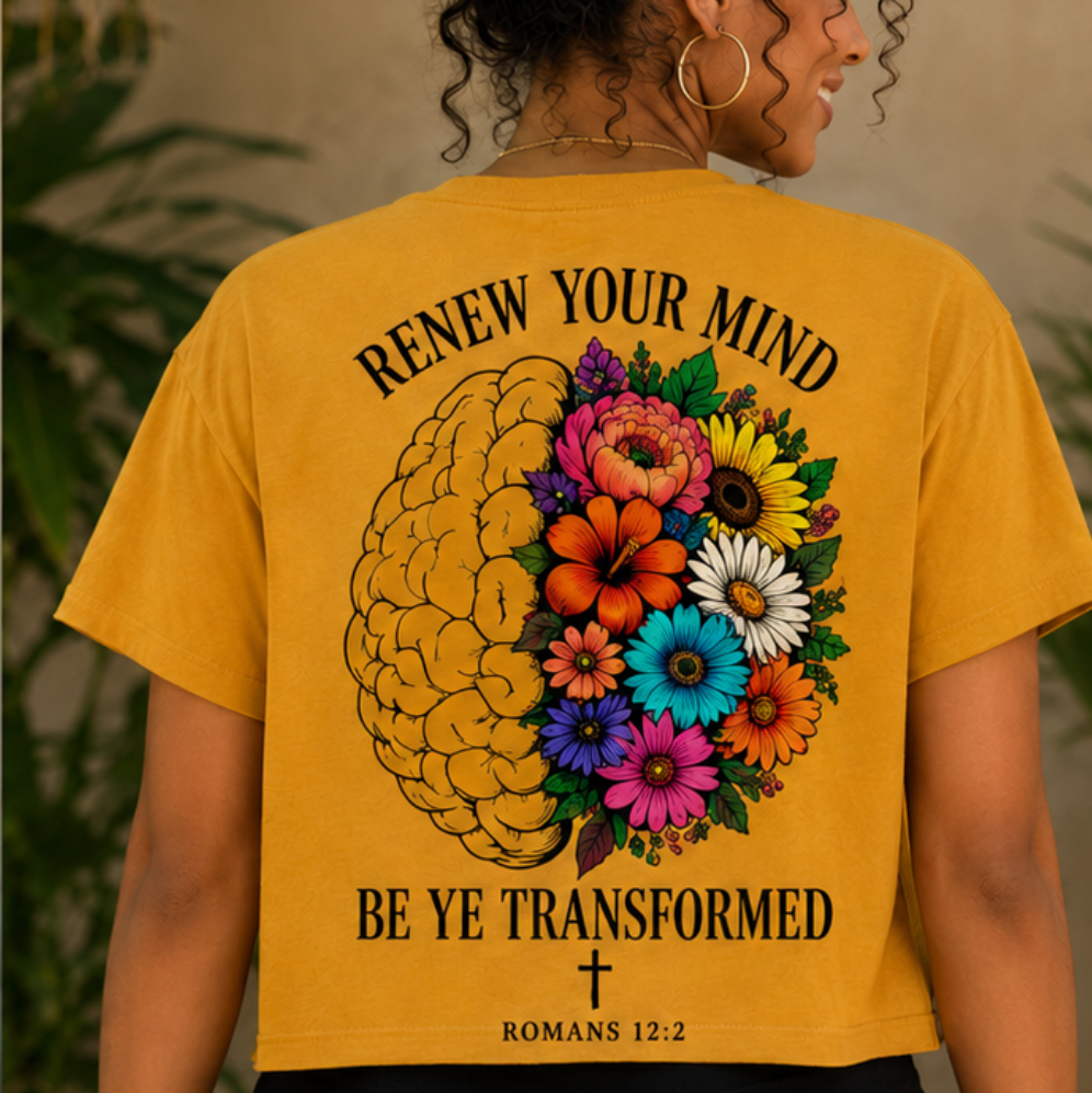 A woman viewed from behind wearing a mustard yellow graphic T-shirt. The back of the shirt features a large printed design with a stylized brain made of flowers and leaves, surrounded by the words 'RENEW YOUR MIND' at the top and 'BE YE TRANSFORMED' with a cross and 'ROMANS 12:2' at the bottom.