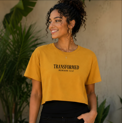 A woman wearing a mustard yellow cropped t-shirt with 'TRANSFORMED ROMANS 12:2' printed in black on the chest, paired with black pants, set against a warm neutral wall with green plants.